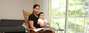 Mother sitting in a wooden rocking chair reading a colorful magazine to a smiling baby on her lap in a bright living room with large windows.
