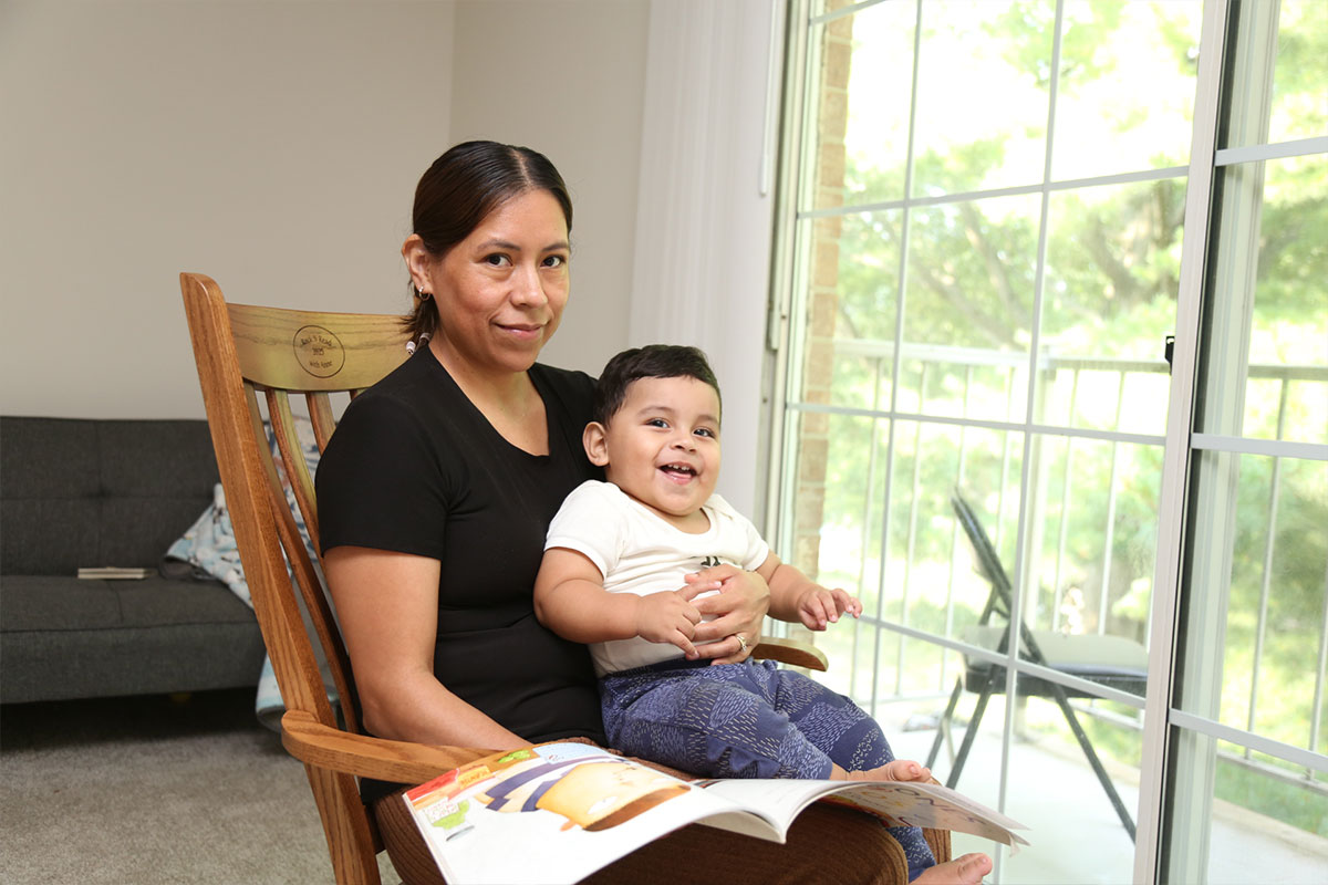 Mother sitting in a wooden rocking chair by a large window, reading a magazine with her smiling toddler on her lap.
