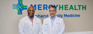 Two doctors in white coats stand side by side, smiling in front of a Mercy Health sign. They’re in a medical office setting.