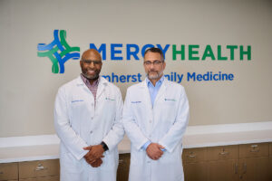 Two doctors in white coats standing in a clinic lobby with a Mercy Health sign in the background, smiling at the camera.