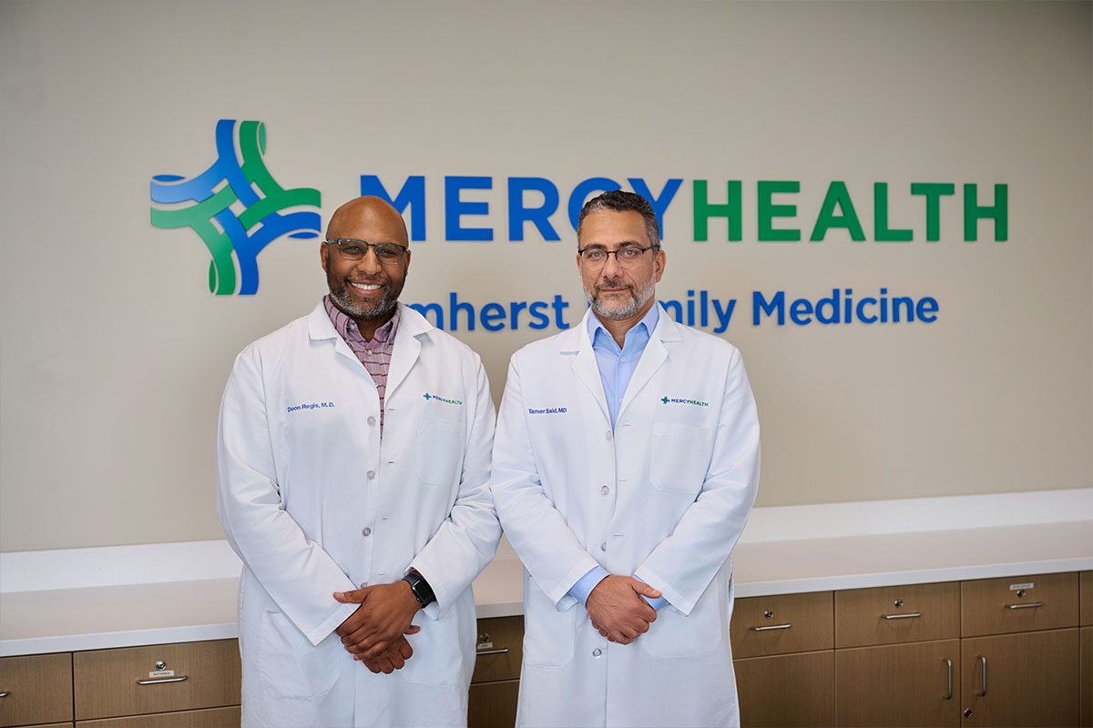 Two doctors in white coats standing in a clinic lobby with a Mercy Health sign in the background, smiling at the camera.
