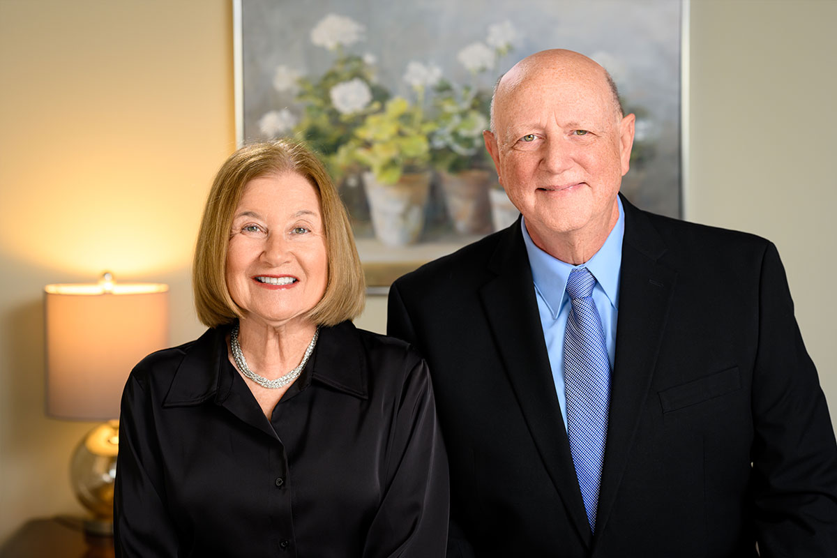 Smiling elderly couple in formal attire posing together in a warmly lit room, woman in black blouse and pearl necklace, man in a dark suit and blue tie.