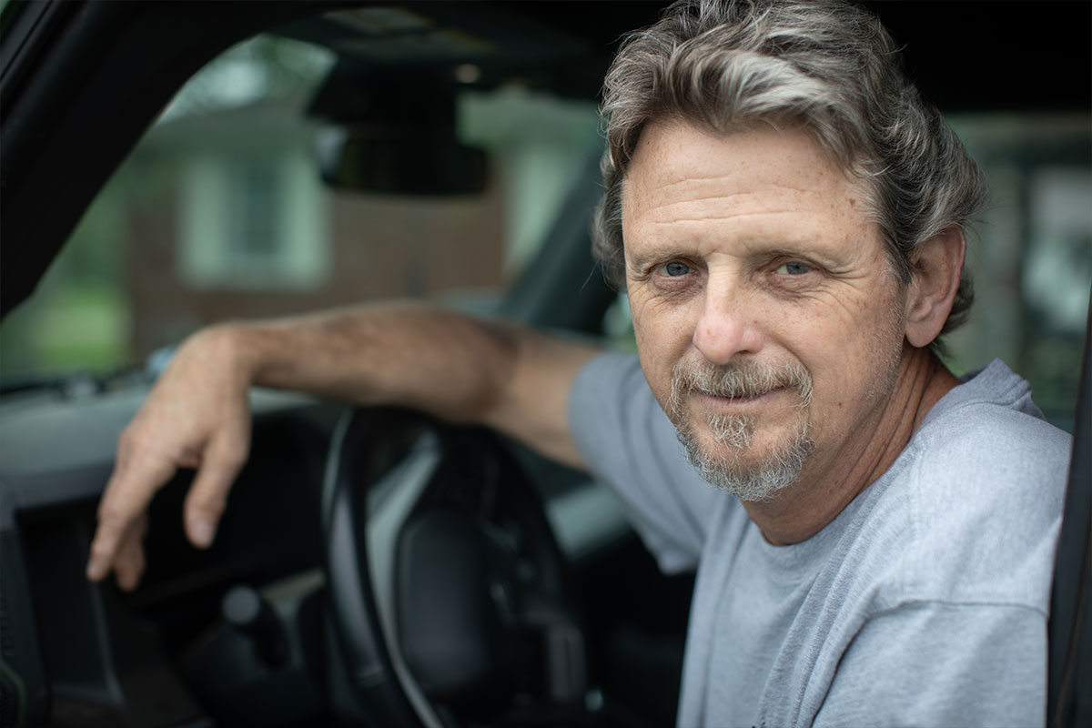 Middle-aged man with gray hair and goatee sits in a car, leaning on the door and looking at the camera.