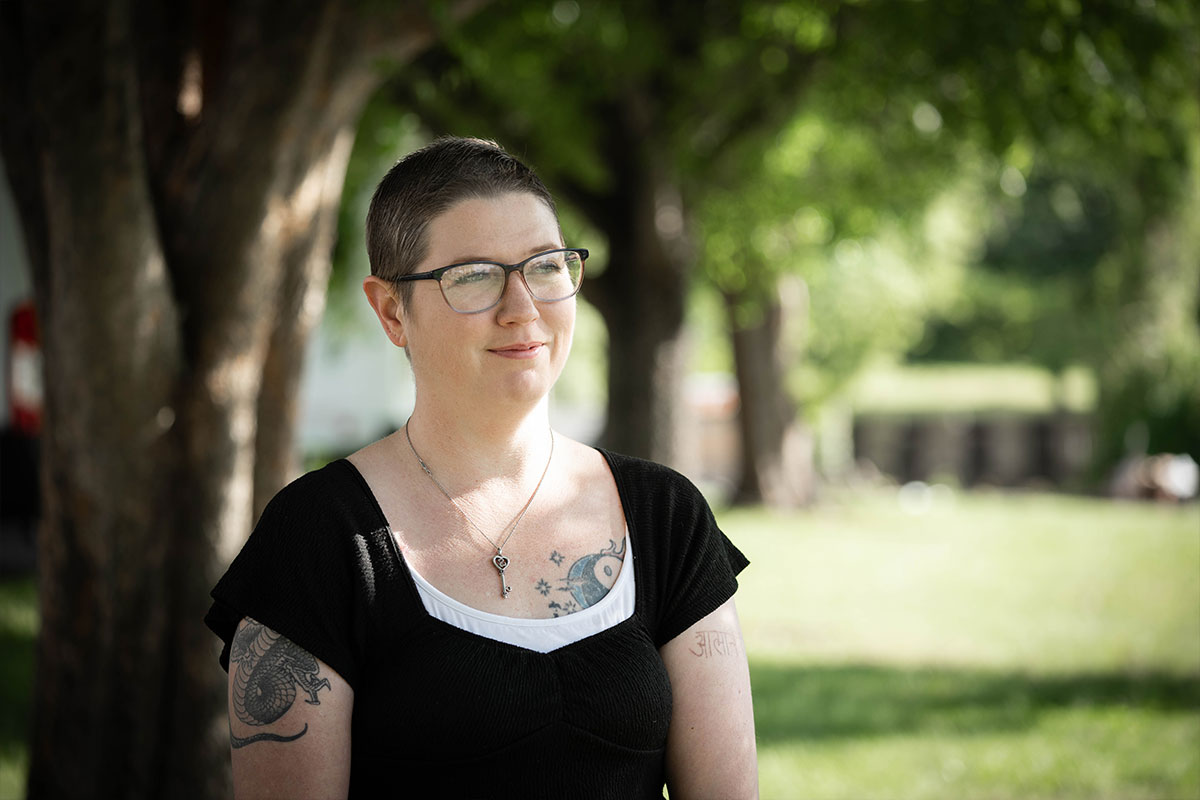 Portrait of a woman with short hair and glasses standing in a sunlit park, wearing a black top and a silver necklace.