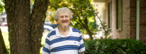 Smiling elderly man with gray hair in a white and blue striped polo shirt standing beside a tree on a sunny residential street.