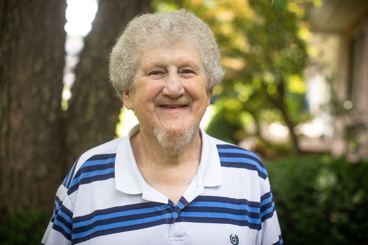 Older man with gray curly hair and goatee smiling outdoors, wearing a white and blue striped polo shirt.
