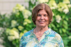 Smiling woman in a blue‑green patterned blouse standing in a garden with greenery behind her.