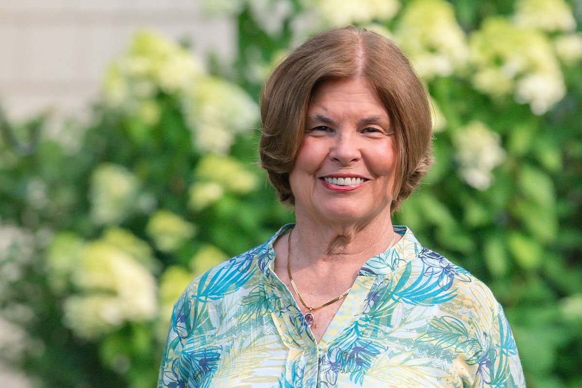 Smiling woman in a blue‑green patterned blouse standing in a garden with greenery behind her.