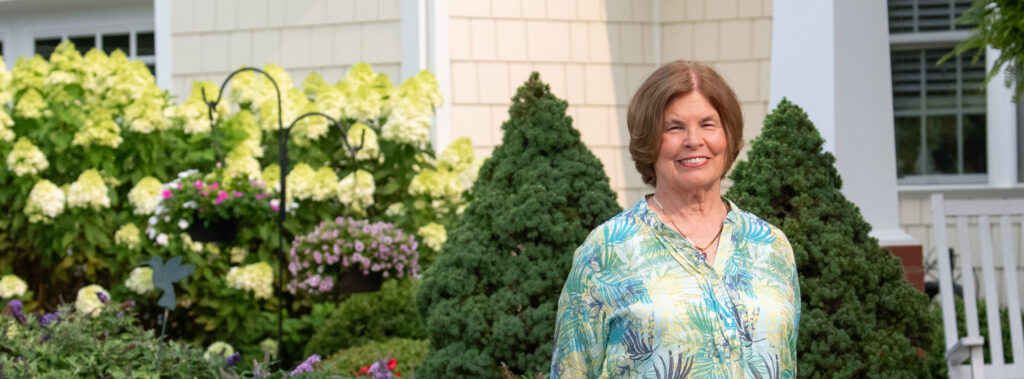 Smiling older woman standing in a garden with yellow hydrangeas, wearing a teal and yellow tropical blouse.