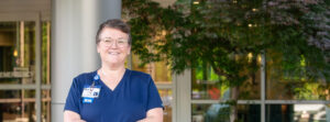 Nurse in blue scrubs smiling outside a hospital entrance, wearing a visible RN badge.