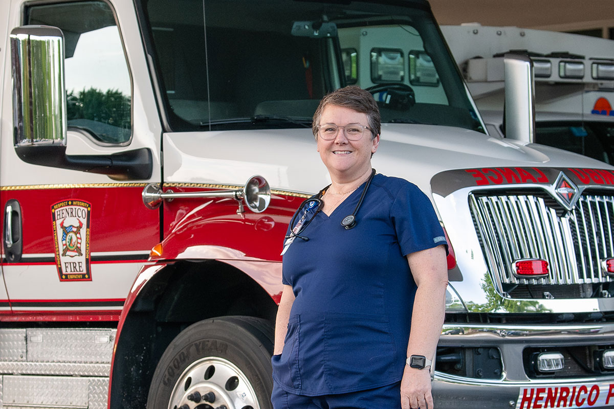 Female healthcare professional in navy scrubs with a stethoscope smiles beside a Henrico Fire Department engine.