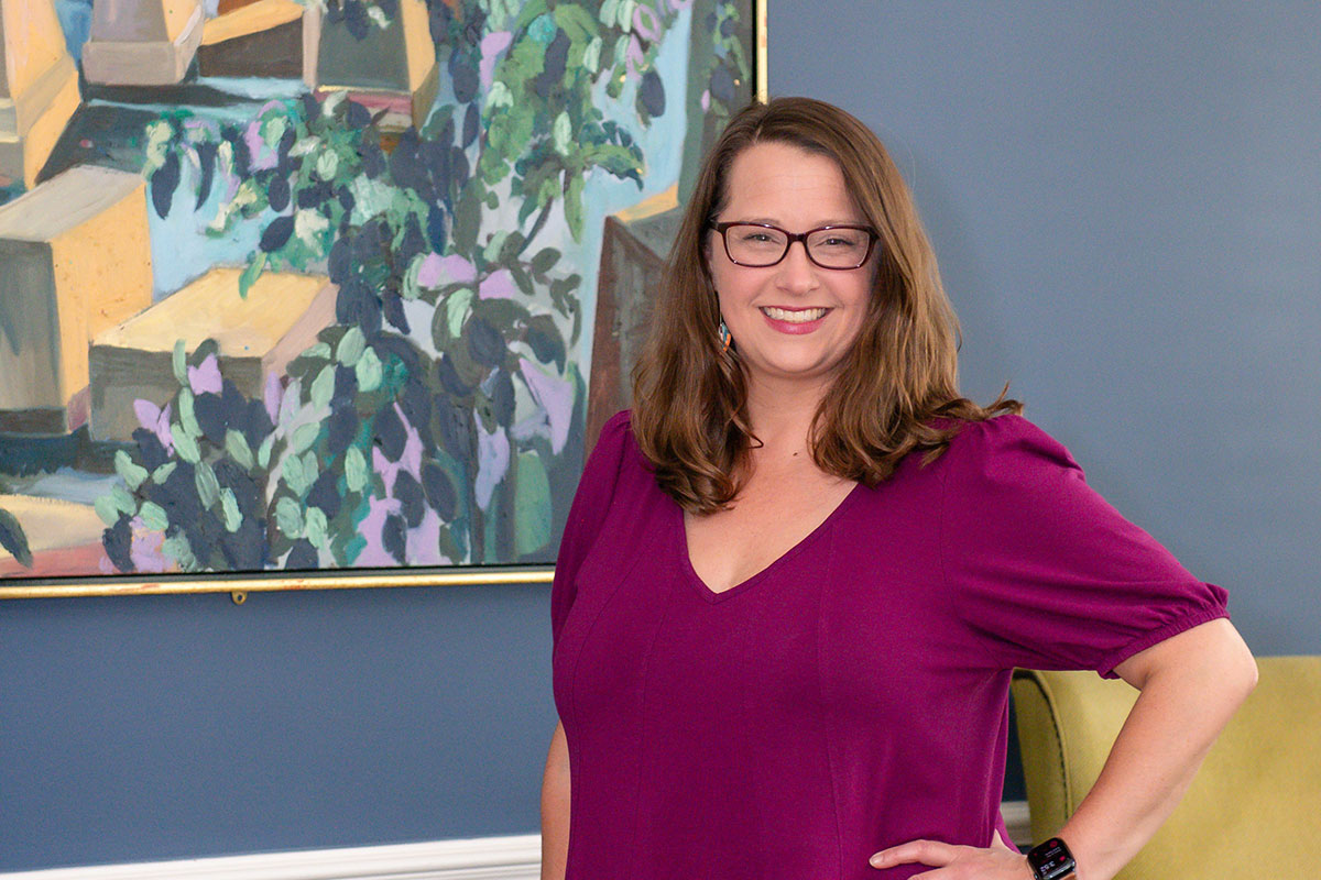 Smiling woman in a magenta blouse with glasses stands with a hand on her hip in front of a blue wall and abstract painting.