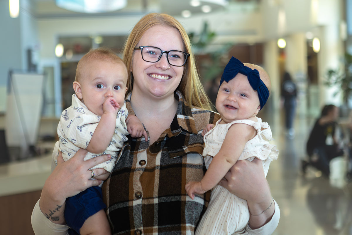 Smiling woman with glasses holding two babies in a bright shopping mall hallway.