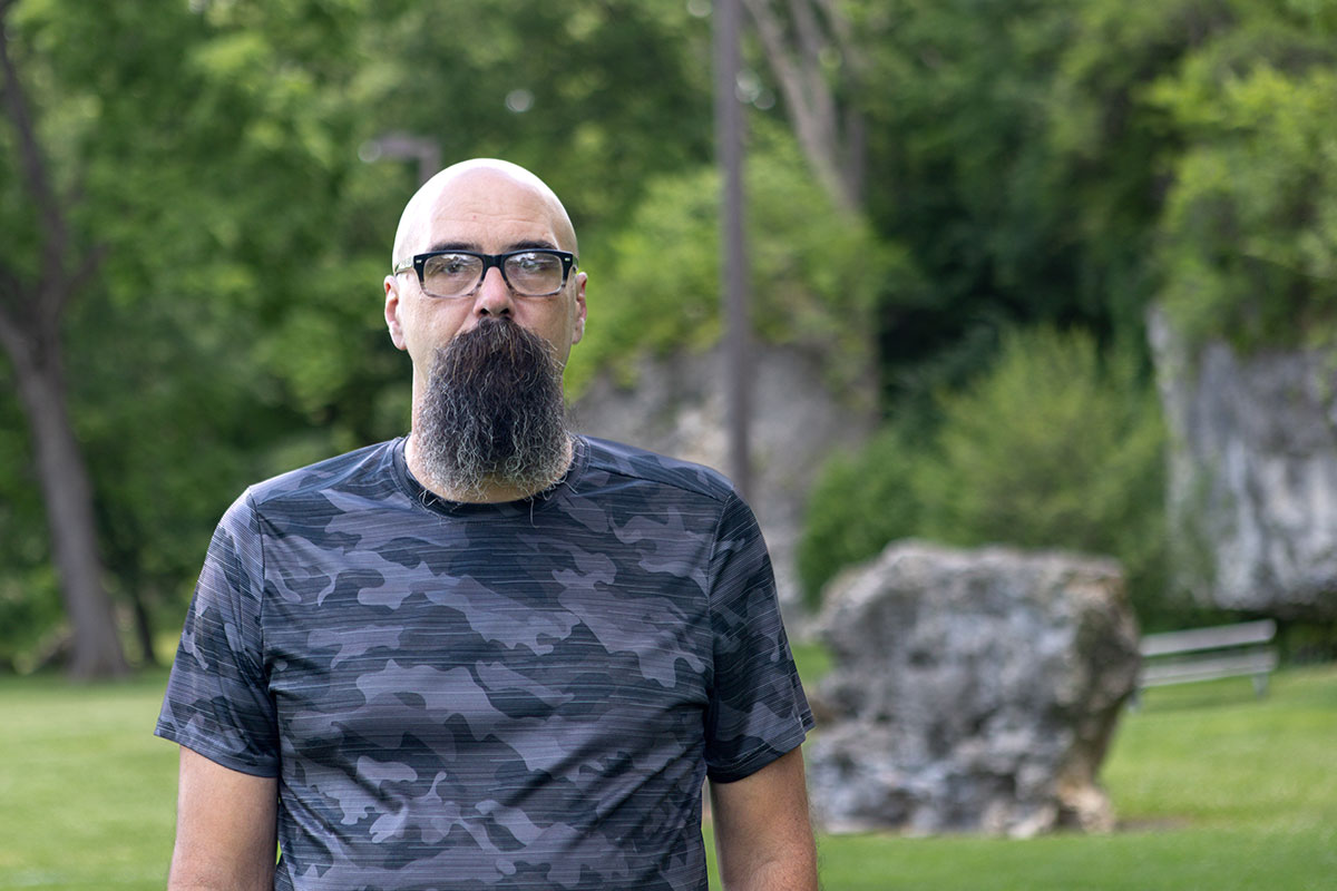 Bald man with glasses and a long gray goatee stands in a park wearing a dark camouflage shirt.