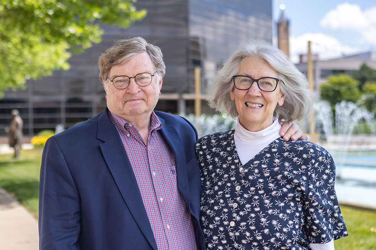 Smiling elderly couple standing outdoors with arm around each other, in front of a modern building and fountain.