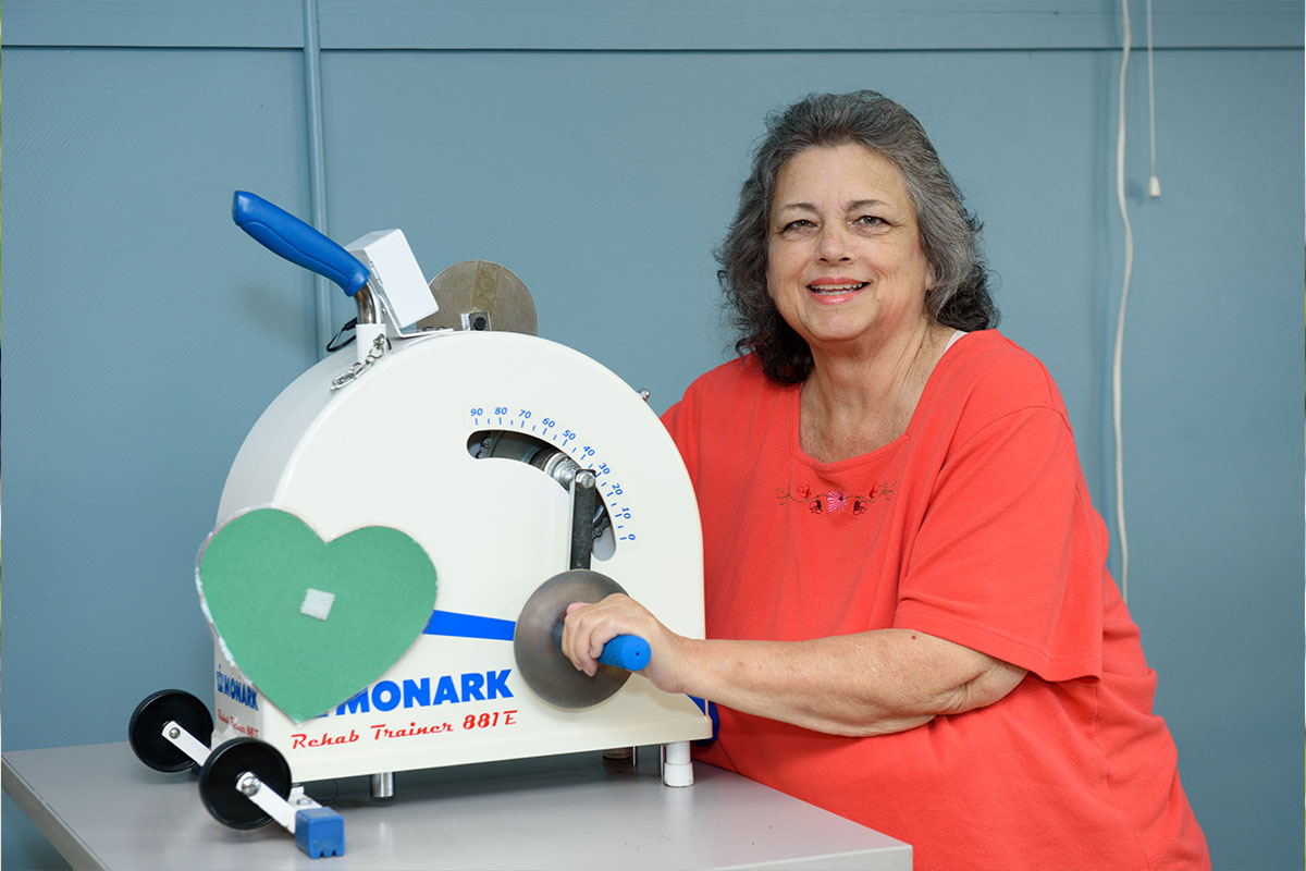 Smiling woman in a coral-red shirt sits at a white rehabilitation device with a blue handle and a large dial keying the exercise motion.