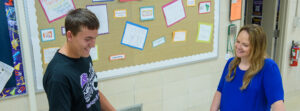 Two people smile and chat in a school hallway beside a bulletin board full of colorful 'Thank You' notes.