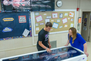 Two students lean over a large interactive touch table displaying a detailed human body diagram.