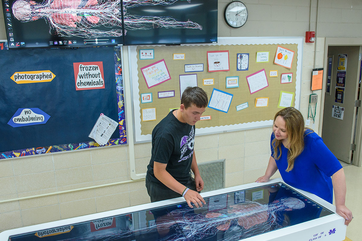Two students lean over a large interactive touch table displaying a detailed human body diagram.