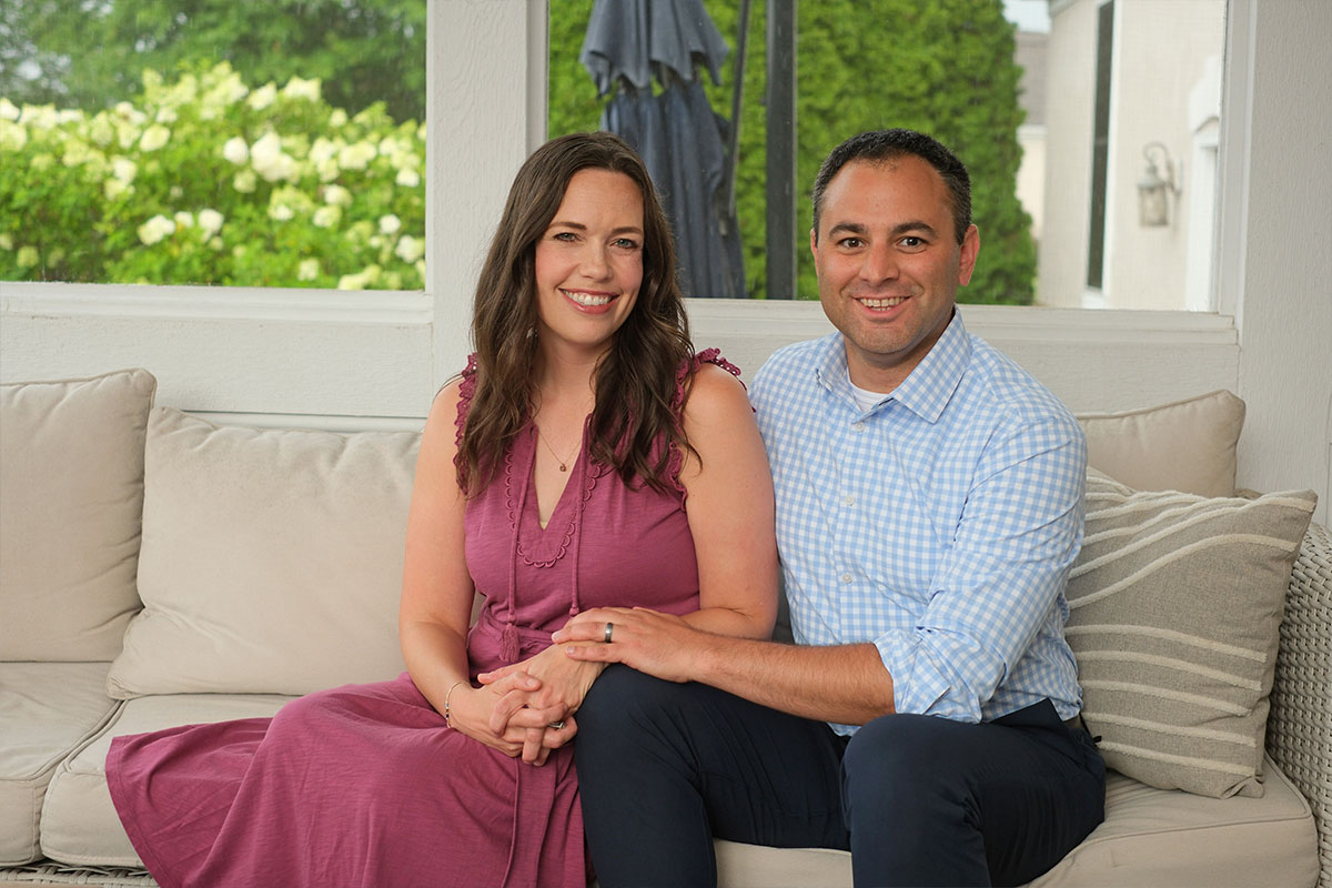 Smiling couple sits on a beige outdoor sofa, holding hands, in a sunlit porch garden setting.
