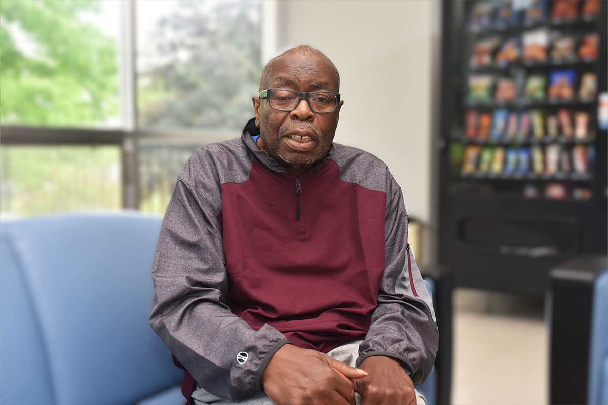 Older man wearing glasses and a maroon-gray jacket sits in a lobby with a vending machine in the blurred background.