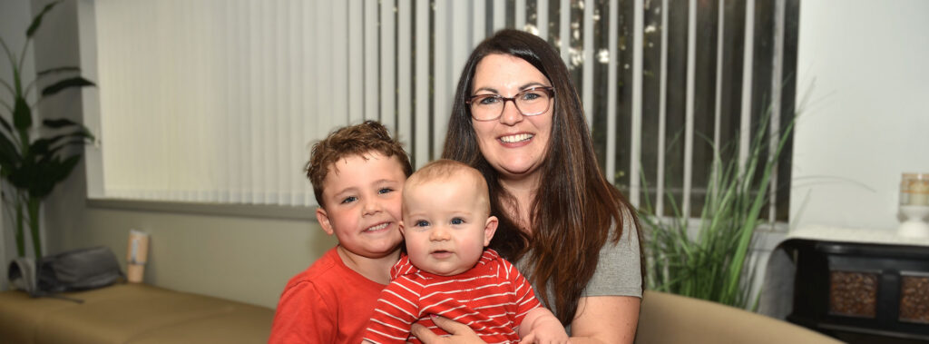 Woman with glasses smiles as she holds a baby while her smiling older son stands beside them on a beige couch in a living room.