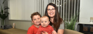 Woman with glasses smiles as she holds a baby while her smiling older son stands beside them on a beige couch in a living room.