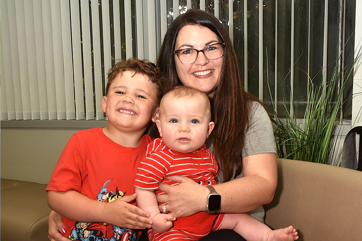 Woman with glasses sits on a beige couch, holding a baby in red stripes while a smiling boy stands beside her.