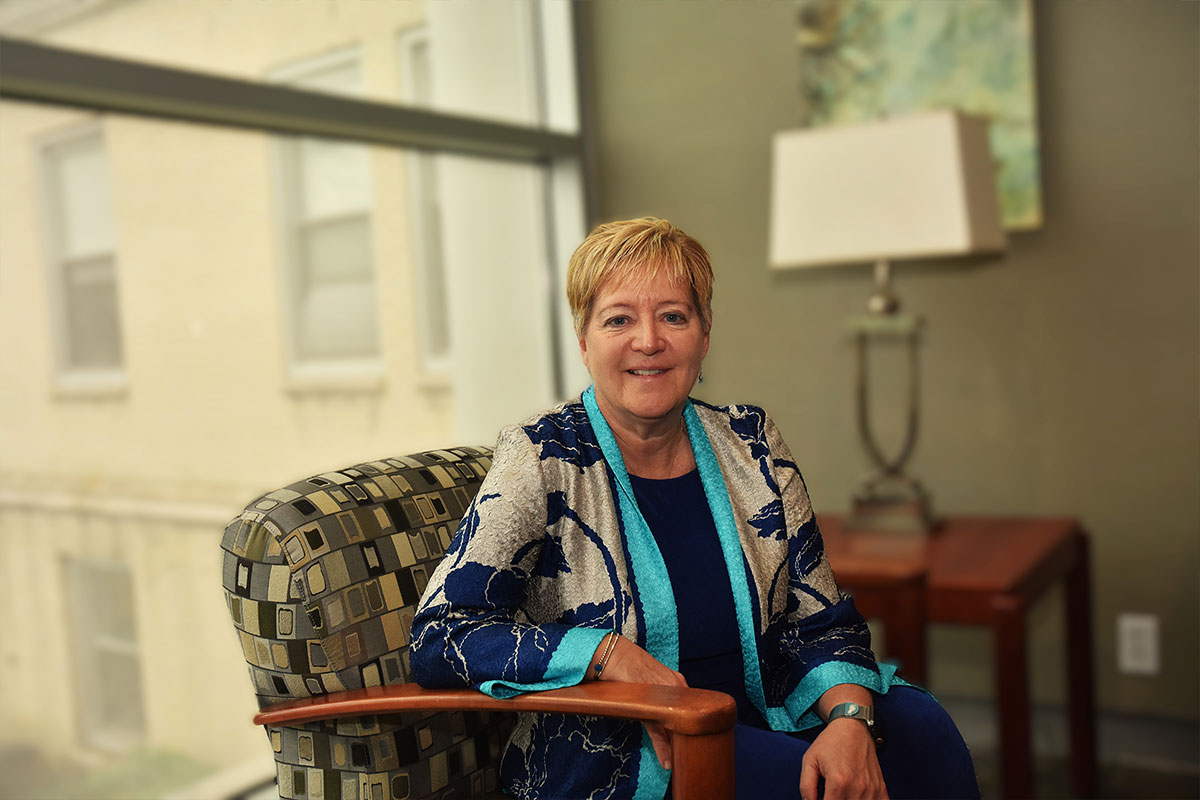 Smiling woman sits in a patterned chair beside a large window in a bright office setting, wearing a blue dress and patterned jacket.