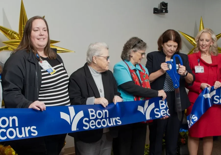 A line of women stand ready to cut a big, blue, ceremonial ribbon with a giant pair of scissors.