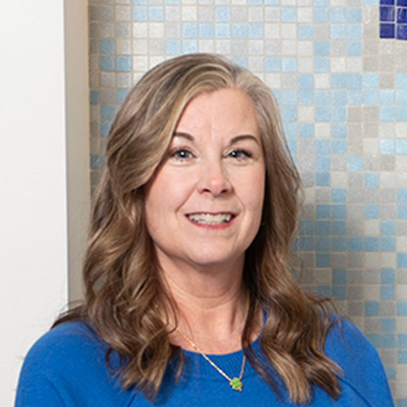 Smiling woman with shoulder-length brown hair in a blue top, standing in front of a blue mosaic tile wall.
