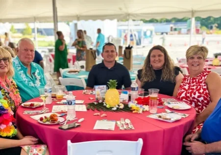 Seven people dressed in floral summer garments sits at an oval table outside, under a tent. The table has a bright red table cloth on it and is laden with beverages and games.