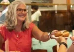 A smiling vendor serves a plate of donuts to a customer at a tasting event.