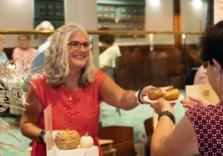 A smiling vendor serves a plate of donuts to a customer at a tasting event.