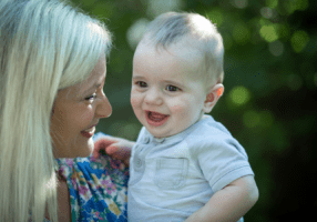 A young, blond mom smiles at her young baby, who she is holding.